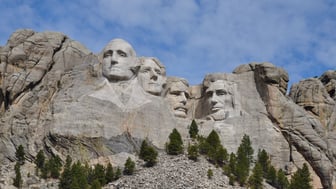 Mount Rushmore's four carved presidential faces in pale granite on a Black Hills cliff, flanked by rough rock outcrops with ponderosa pines on the rubble slope below, under a bright blue sky with thin high clouds.